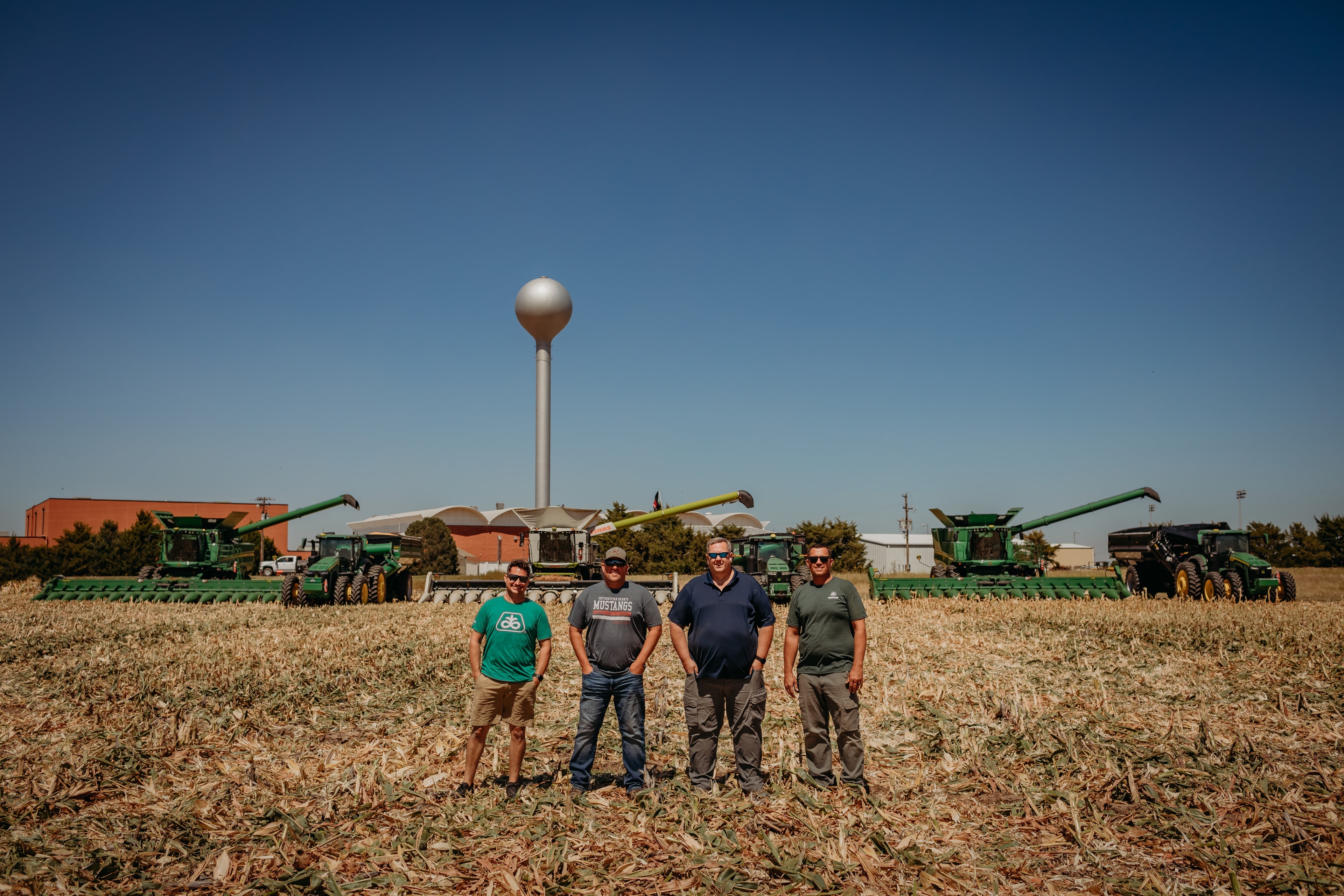 Four foundation members standing in a harvested corn field with John Deere combines and a water tower in the background
